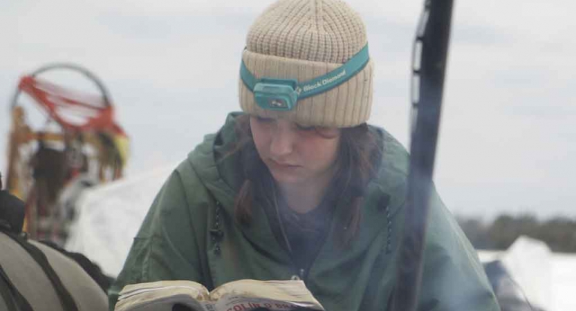 a student wearing winter clothing reads from a book while sitting amongst a snowy landscape